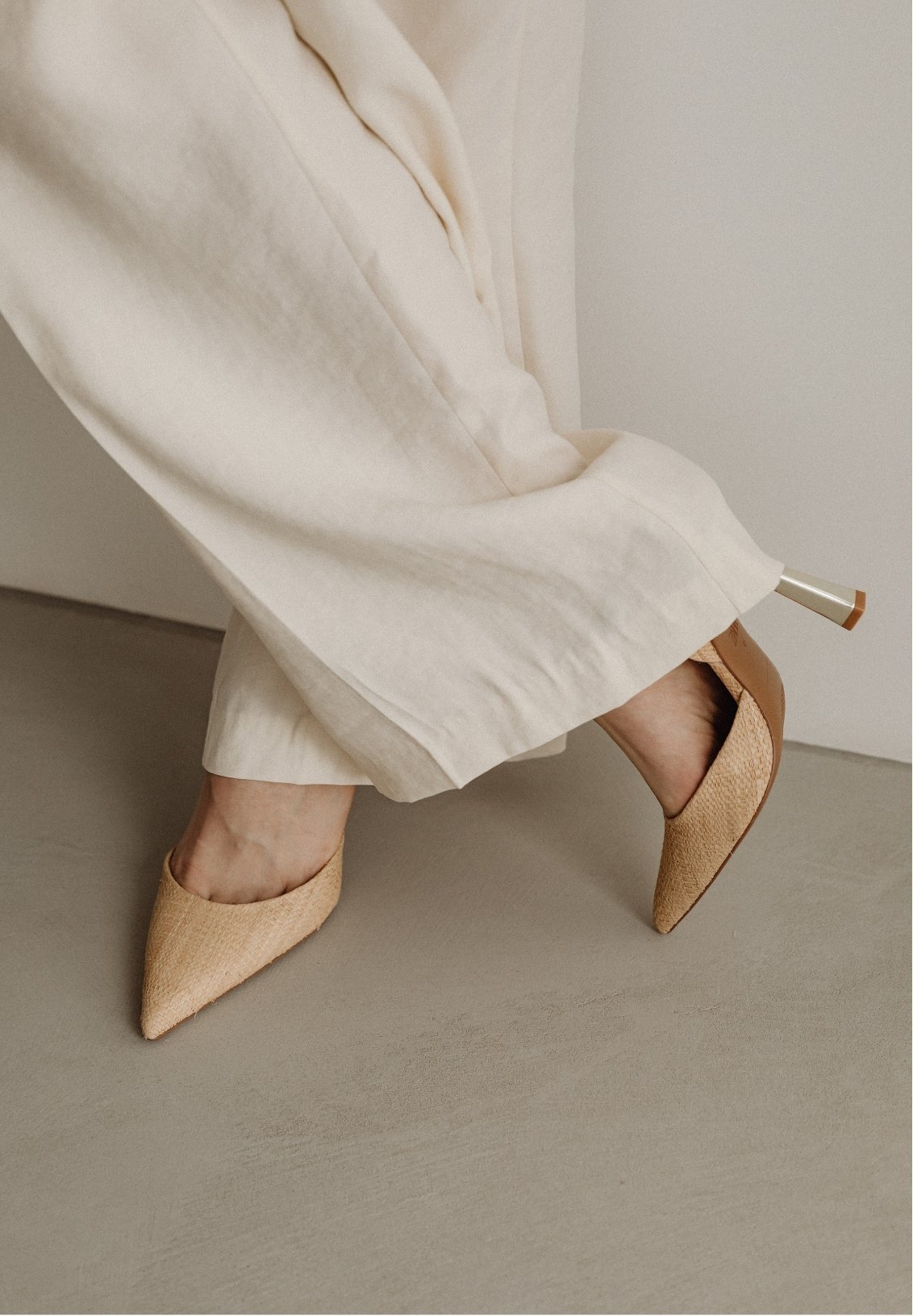 Person wearing beige pointed high heel shoes and cream-colored pants, standing on a smooth light gray surface next to a white wall.