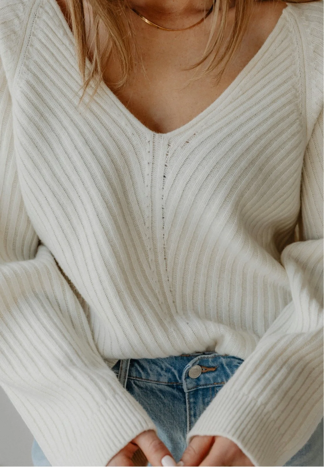Close-up of a woman wearing a cream-colored ribbed knit sweater with a V-neck, partially showing a gold necklace, and denim jeans.