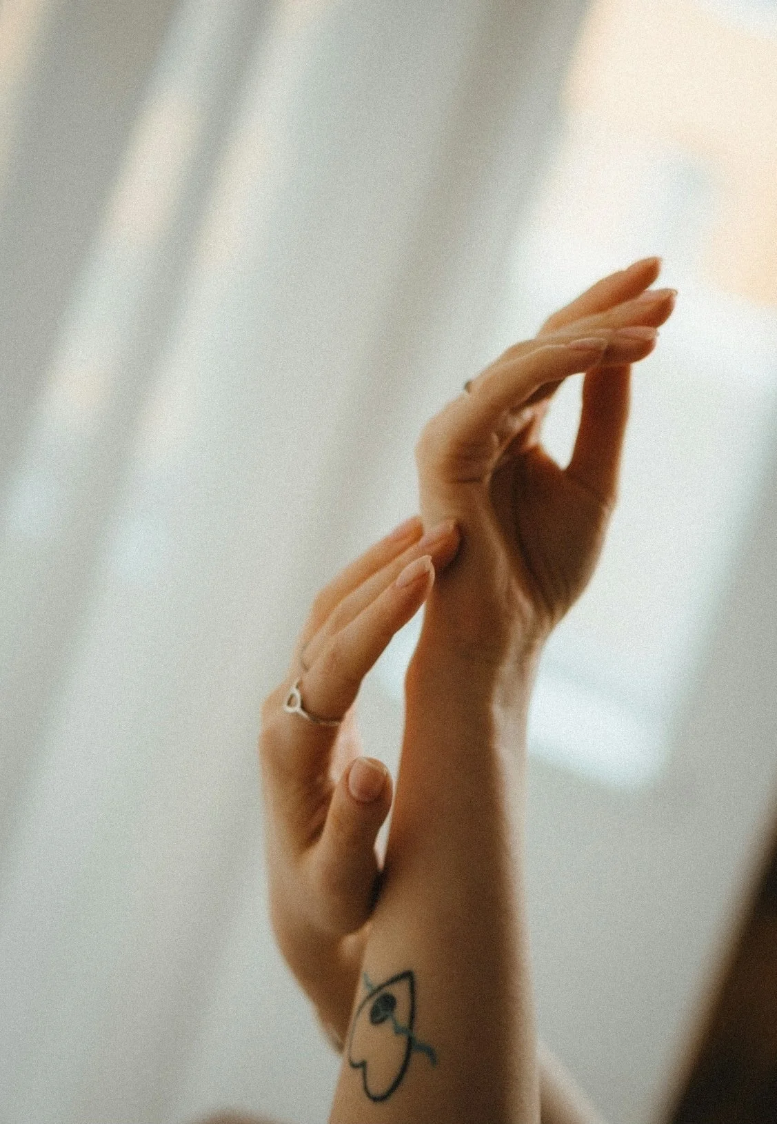 Close-up of two hands with fingers touching, skin tones are light, with a small black and white tattoo of an eye on the wrist, and a simple silver ring on one finger. The background is blurred and neutral.