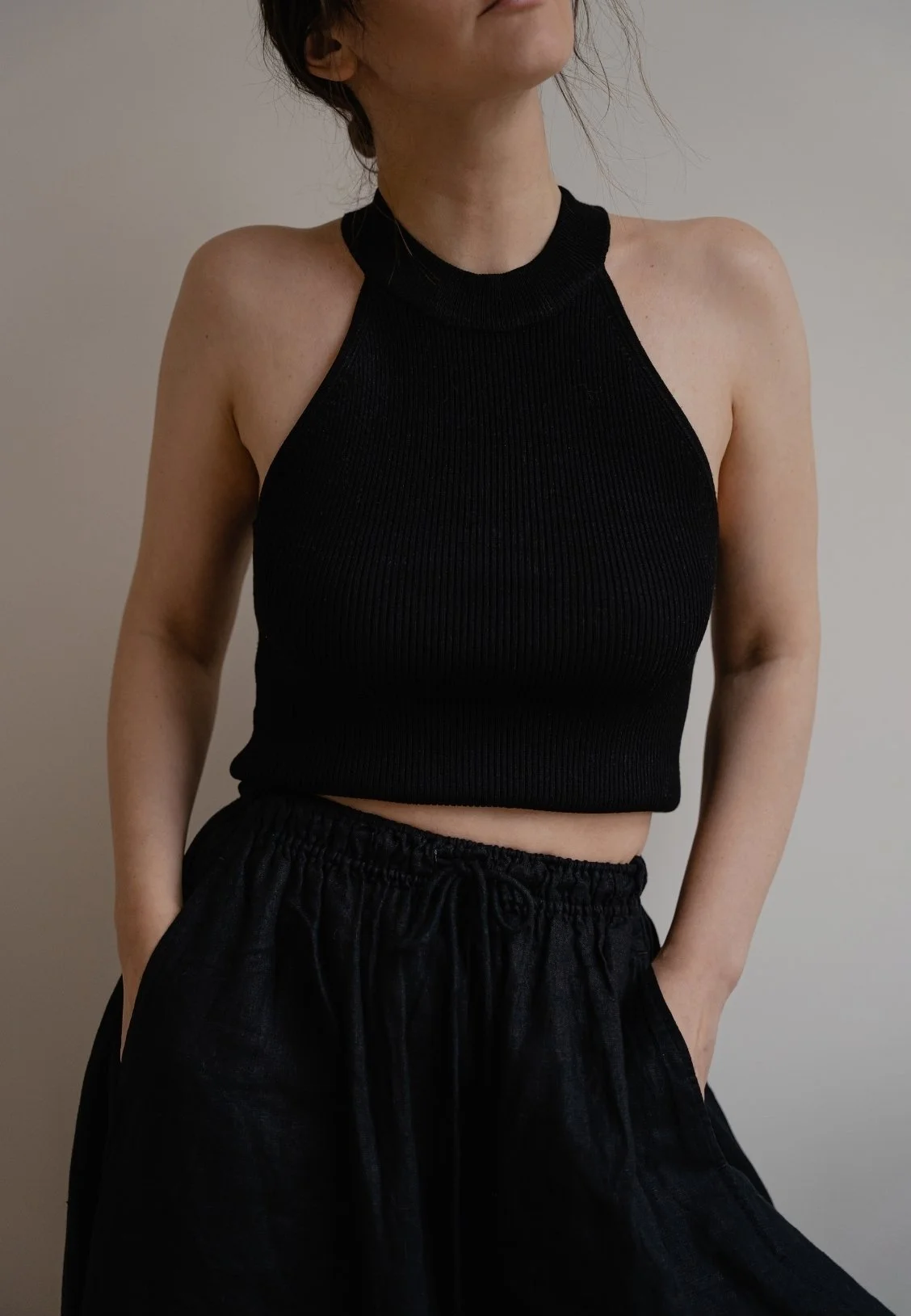 A woman wearing a black sleeveless crop top and loose black pants, standing against a plain gray background.