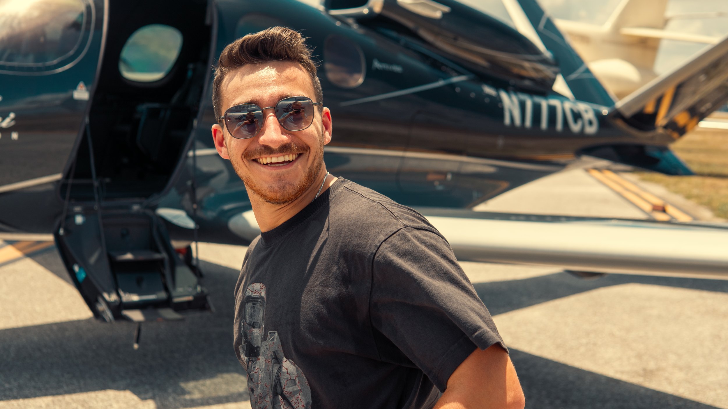 A smiling man with sunglasses standing in front of a black private jet, with the jet's open door visible behind him.
