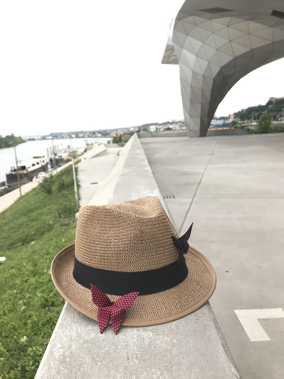 Un chapeau de paille beige avec un ruban noir et deux nœuds rouges avec des pois noirs, placé sur un parapet en béton au bord d'un quai ou d'une promenade avec un bâtiment futuriste en forme d'arche en arrière-plan.