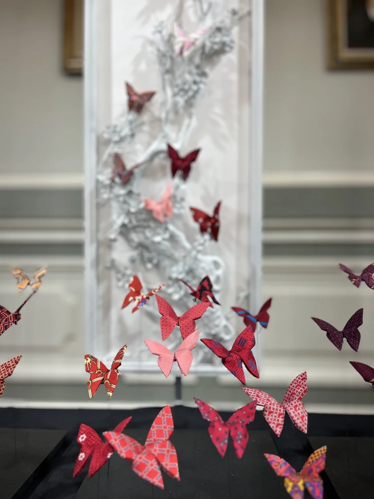 Installation artistique avec des papillons en papier colorés suspendus en vol devant une sculpture blanche d'arbre.