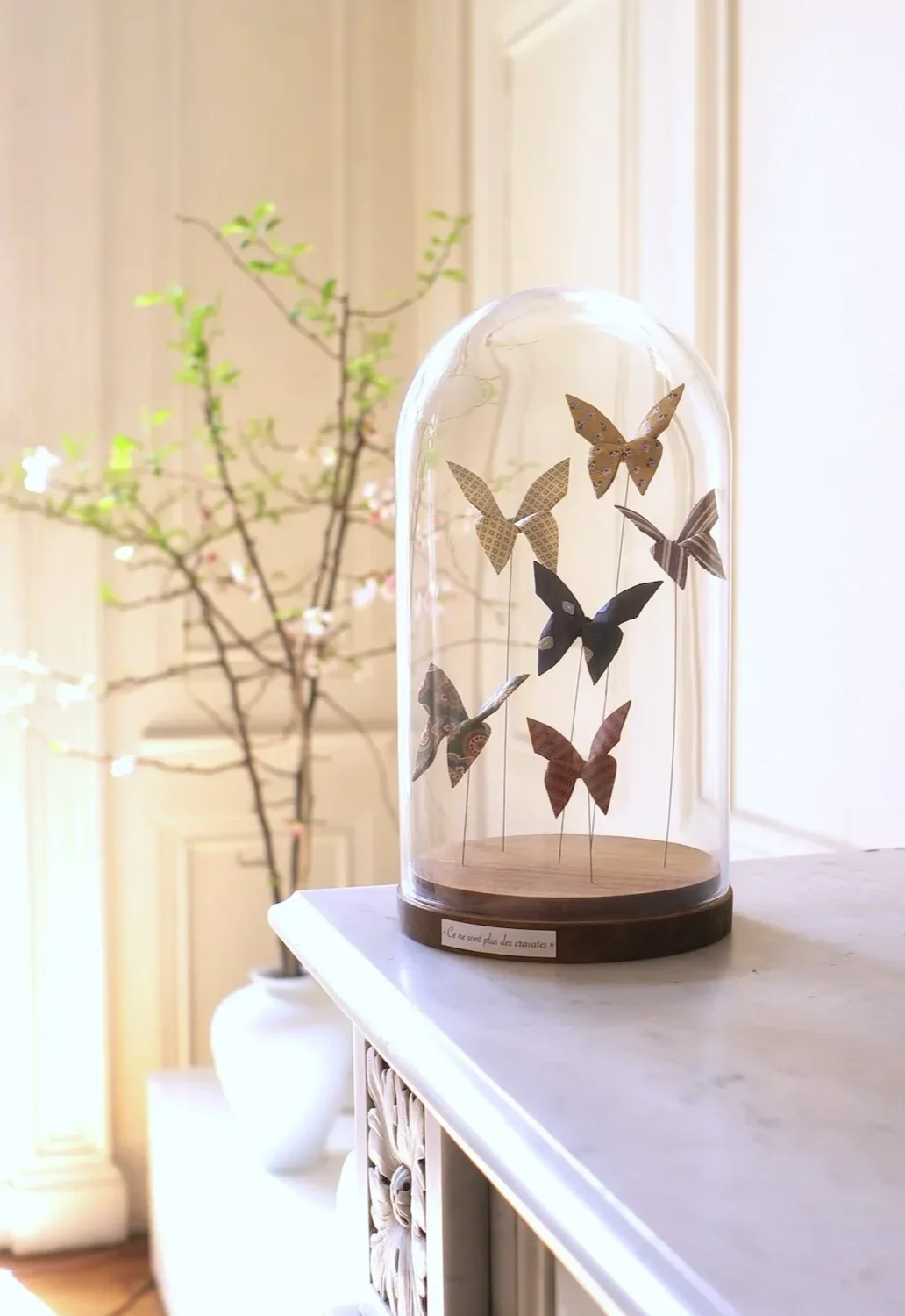 Une décoration artistique avec des papillons en papier suspendus dans une cloche en verre posée sur une table en bois, avec un fond de fenêtre et un arbre décoratif avec des feuilles en arrière-plan.