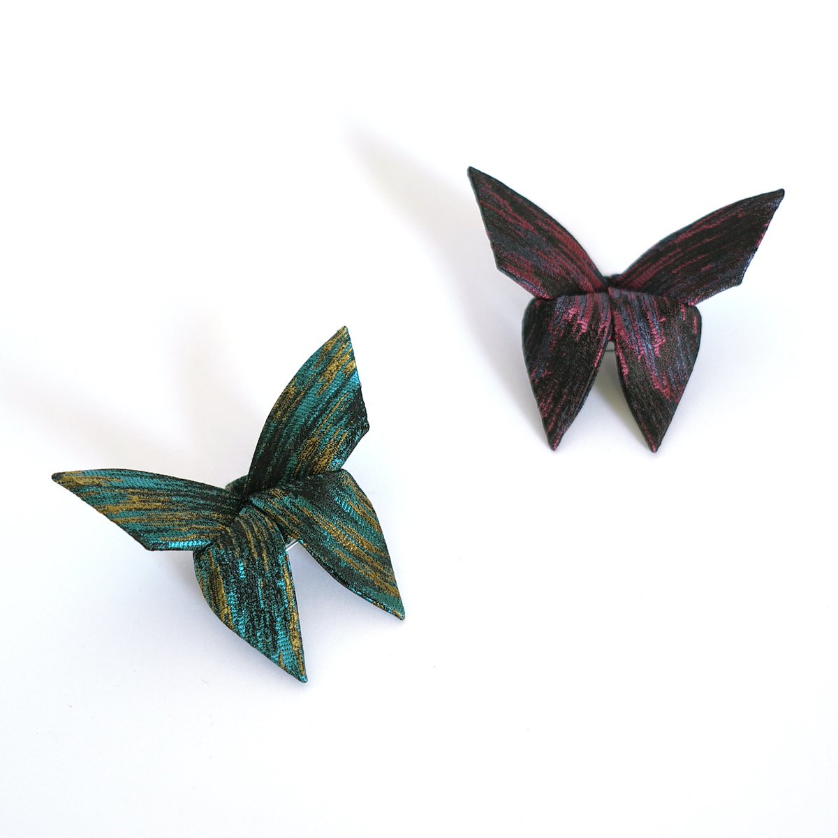Deux pinces en forme de papillons en tissu coloré, un vert avec des reflets dorés et un rouge foncé avec des reflets violets, posées sur un fond blanc.