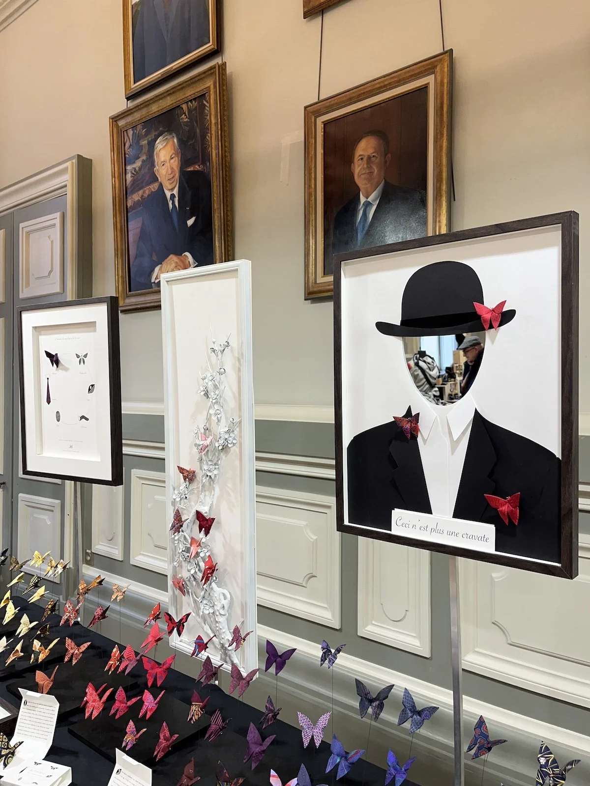 Une exposition de portraits de silhouettes d'hommes avec des cadres dorés, des œuvres d'art de papillons colorés sur un mur blanc, et une collection de papillons en origamis de différentes couleurs sur une table. Un panneau indique en français : "Ceci n'est plus une cravate."