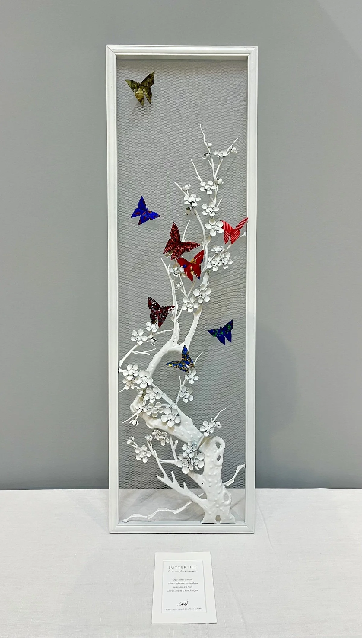 Une œuvre d'art représentant un arbre blanc avec des branches et des fleurs, décorée de papillons colorés en papier suspendus autour, exposée dans une vitrine blanche.
