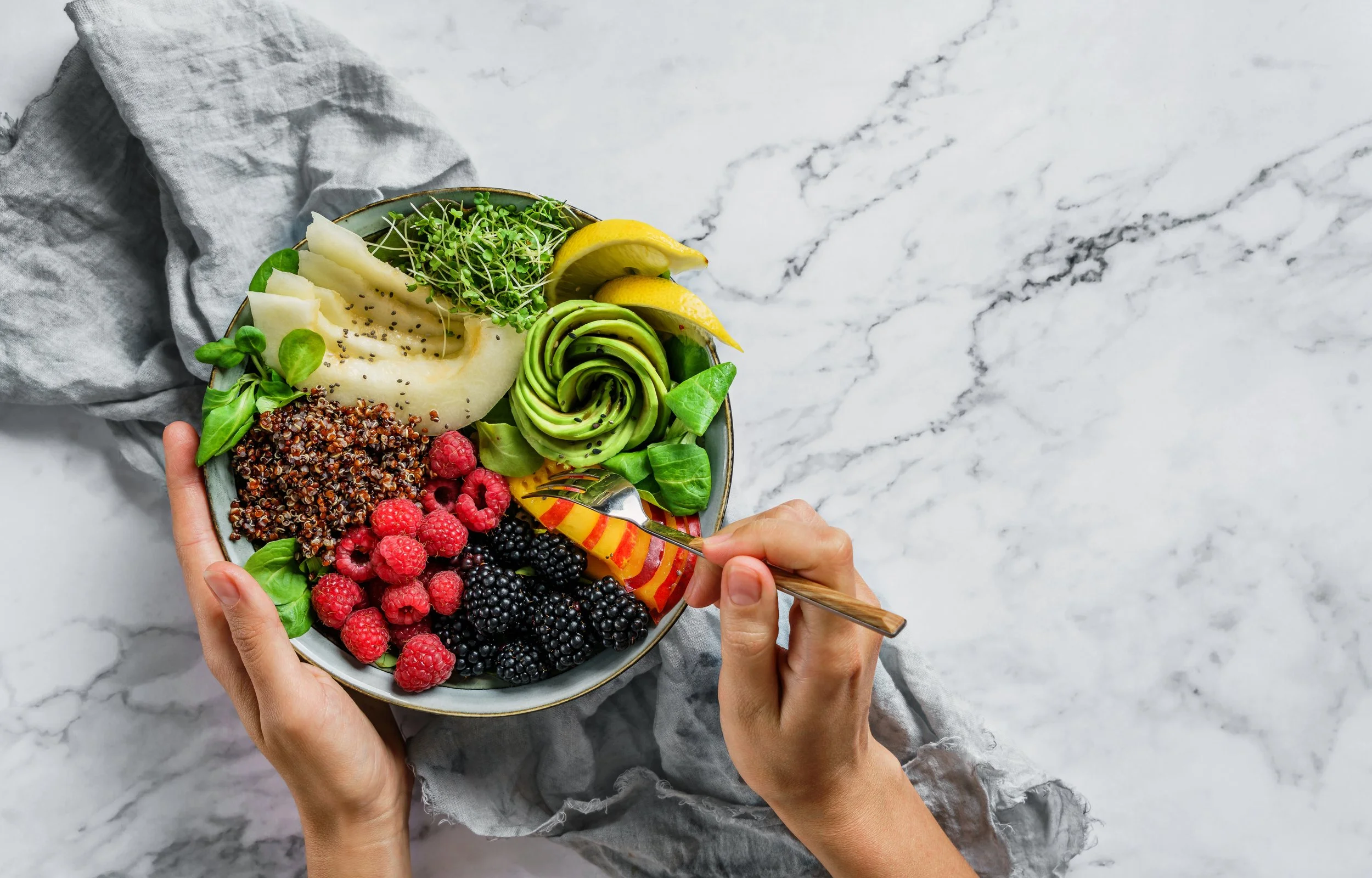 Hands holding a bowl of colorful berry and fruit salad on a white marble surface.