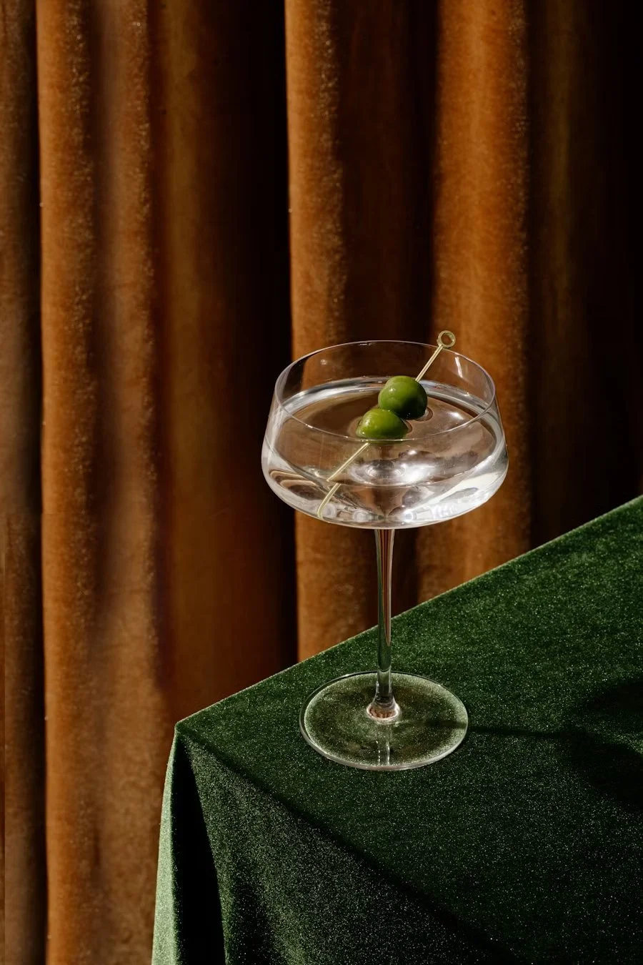 Martini with olives in a coupe glass on a green velvet table against a rich brown curtain backdrop.
