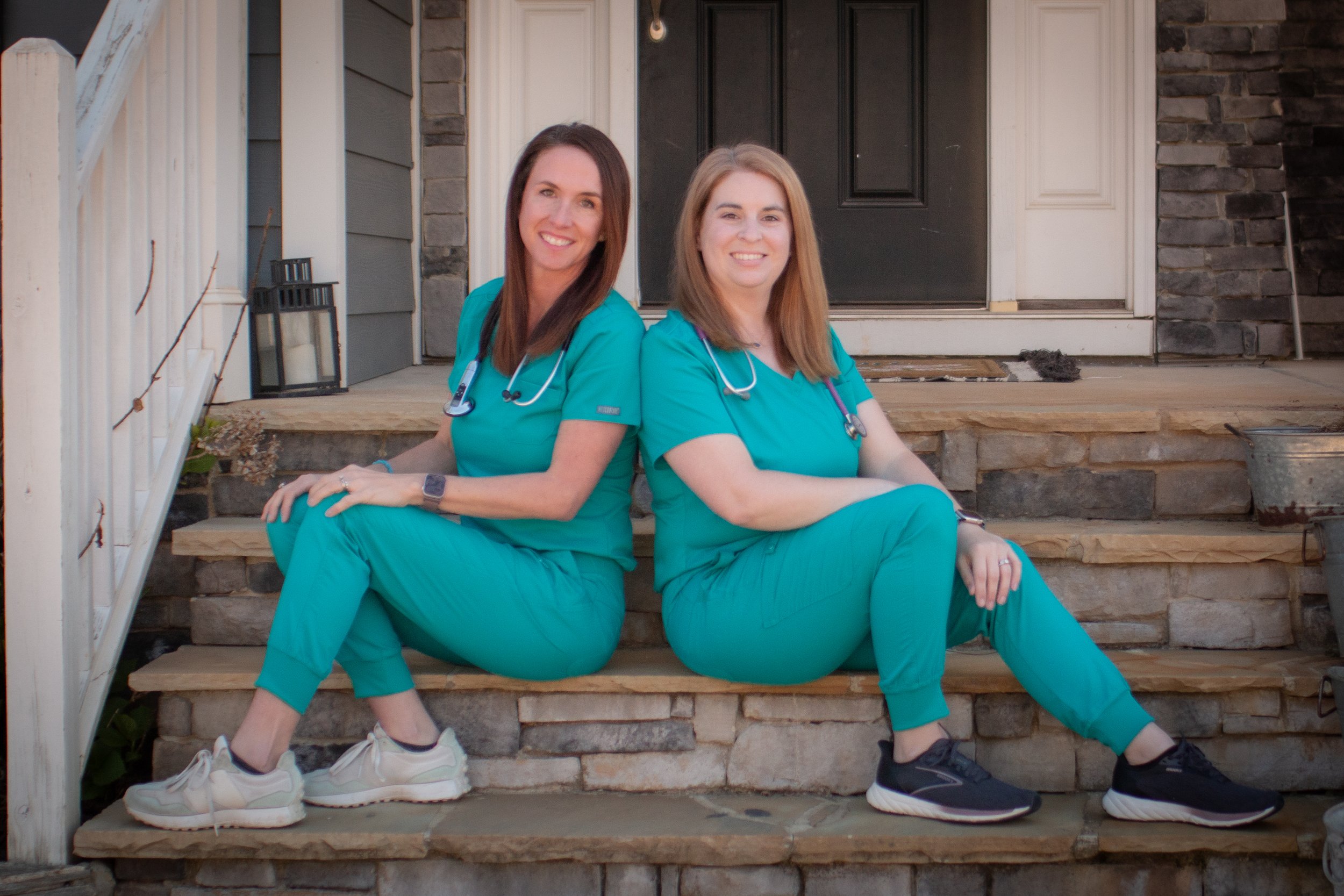 Two women wearing teal medical scrubs sitting on the front steps of a house, smiling at the camera. They have stethoscopes around their necks, and both are wearing athletic shoes.