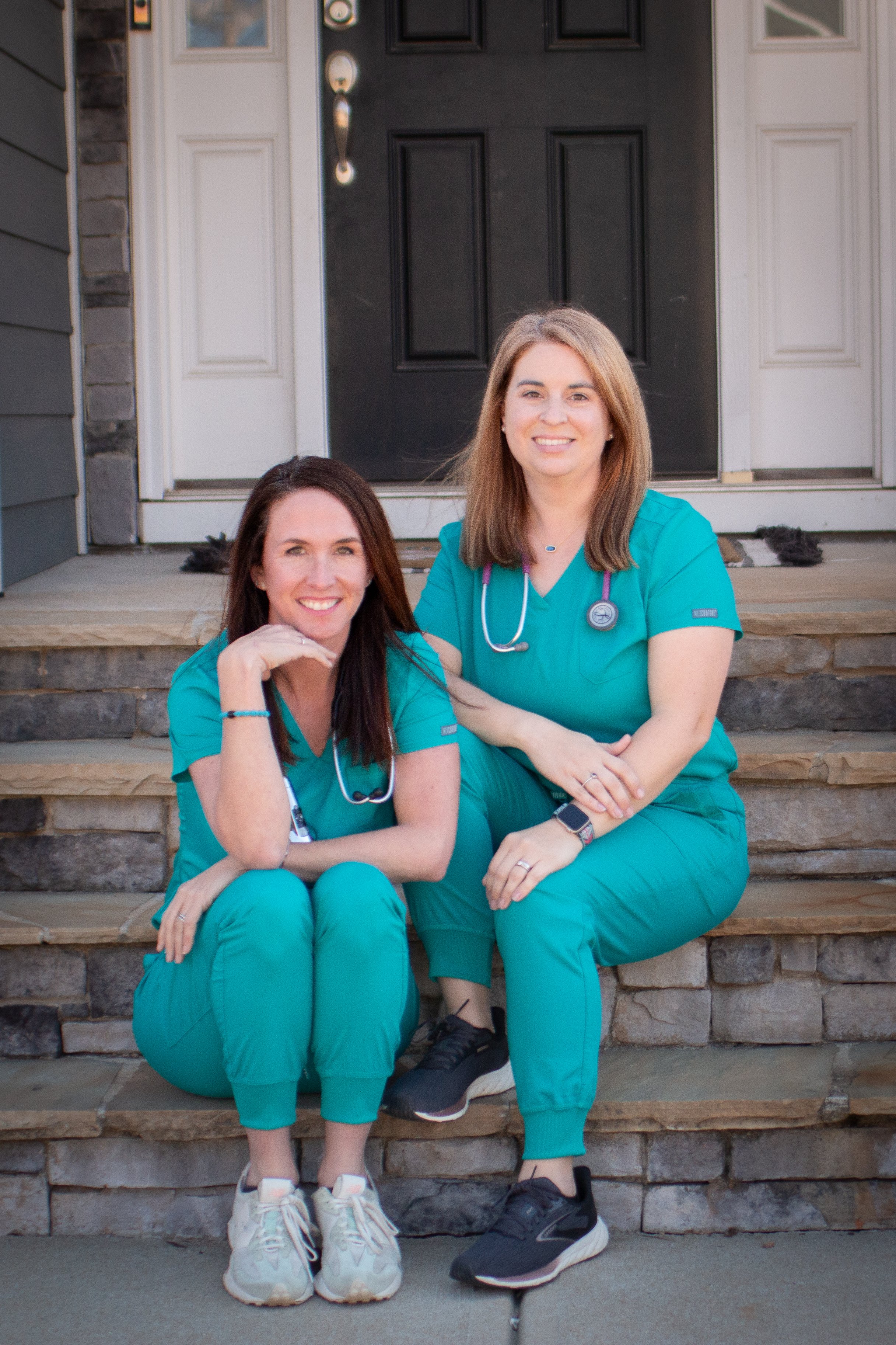 Two women in teal medical scrubs sitting on stone steps outside a house, smiling at the camera.