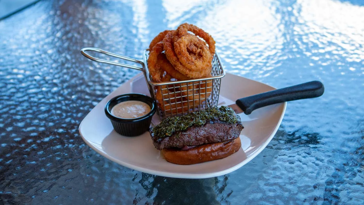 Fried onion rings, steak with chimichurri sauce, and fried potato on a white plate, with dipping sauce, on a glass table.