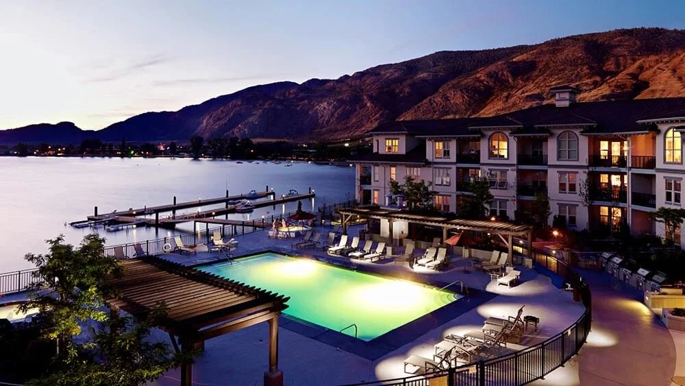 Luxury lakeside hotel at dusk featuring a swimming pool with lighting, a dock with boats, and mountain backdrop.