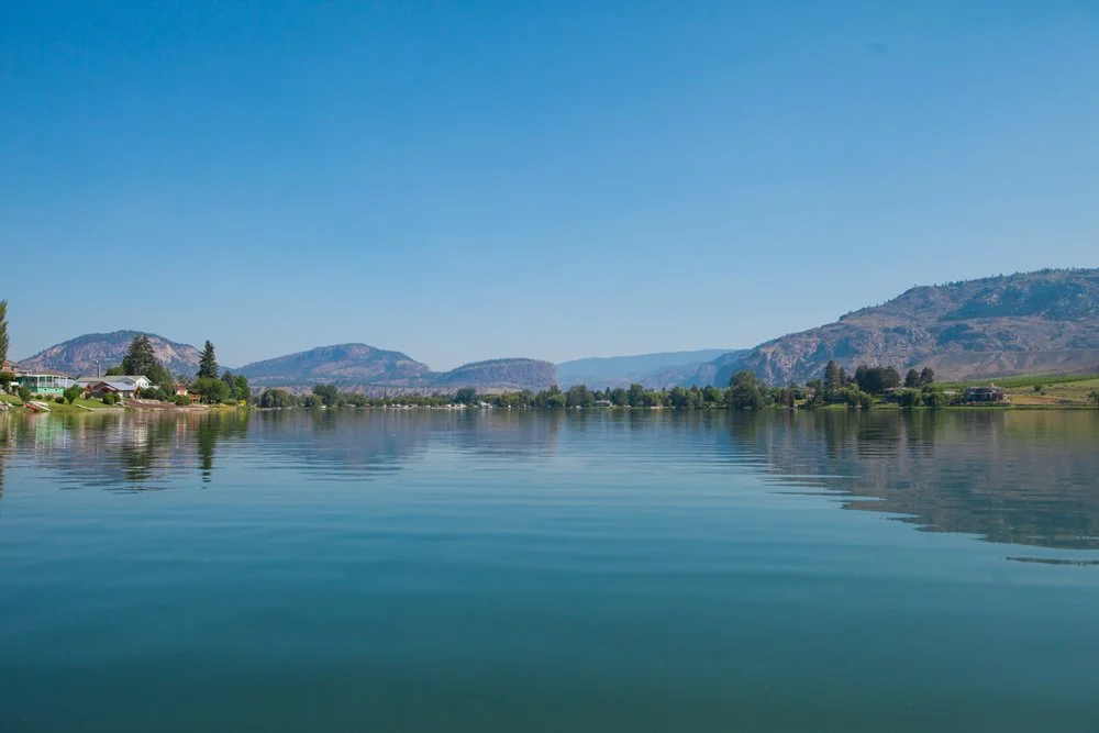 Calm river with houses on the left and mountains in the background under a clear blue sky.