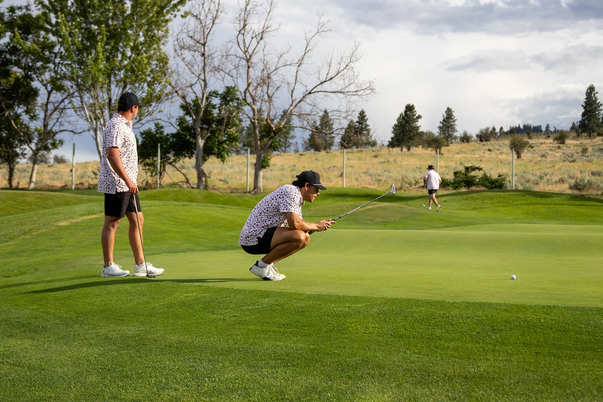 Three men playing golf on a lush green course at Fairview Mountain Golf Club, with one man crouched down preparing to putt, a woman walking in the background, and trees and cloudy sky in the distance.