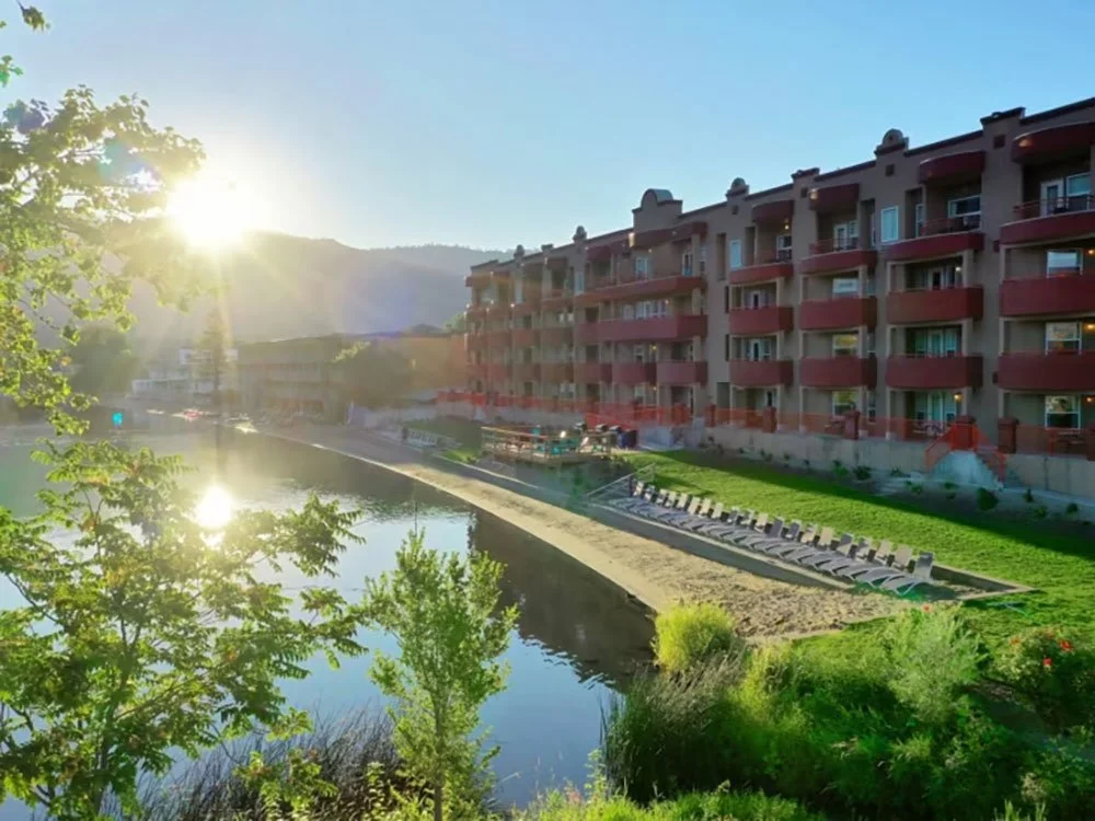 Sunrise over an apartment complex next to a river, with greenery and lounge chairs on a lawn.