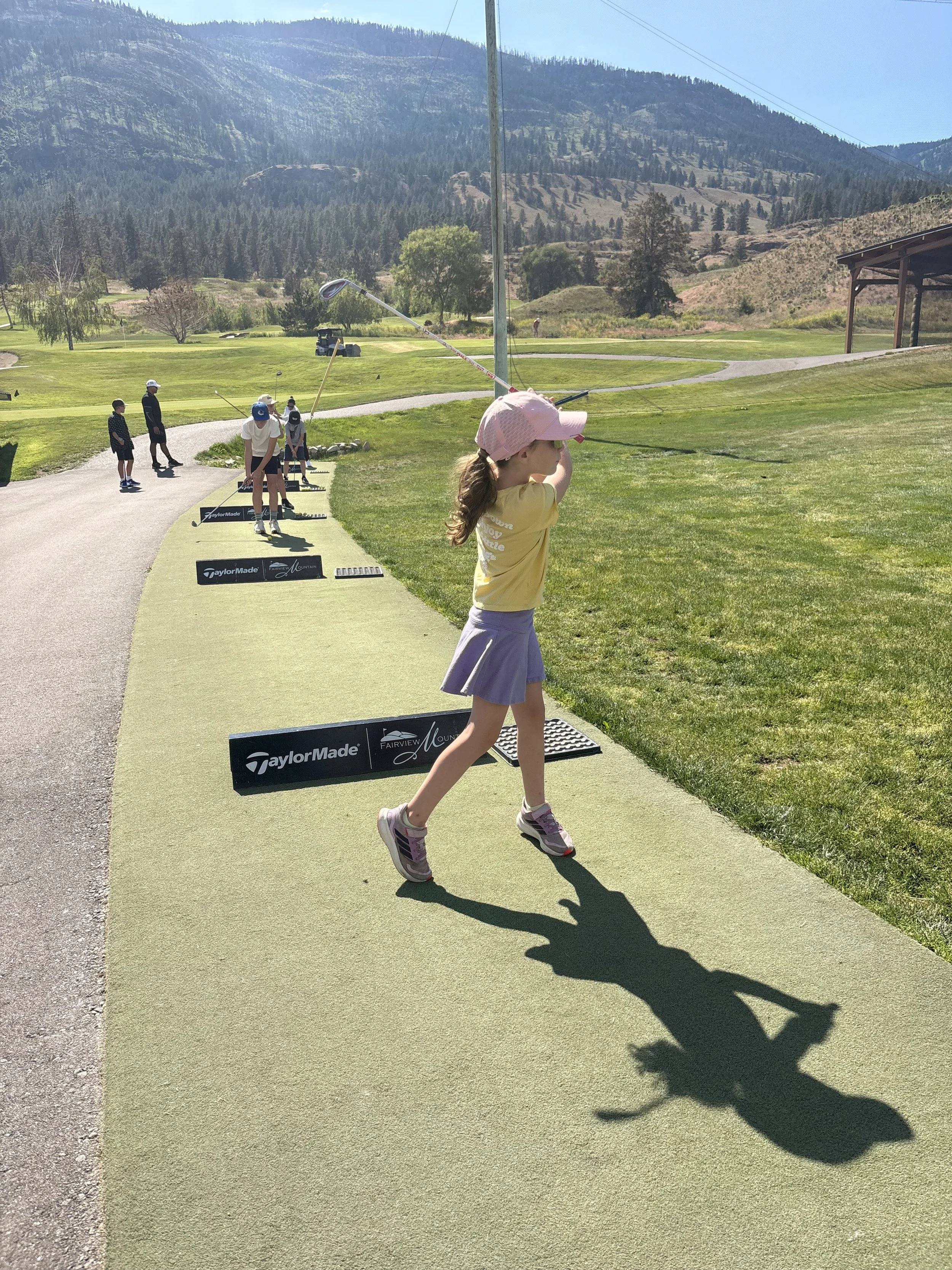 A young girl in a pink hat and yellow shirt practicing golf swings on the Fairview Mountain Golf Club driving range with other children and adults in the background, set against a mountainous landscape.