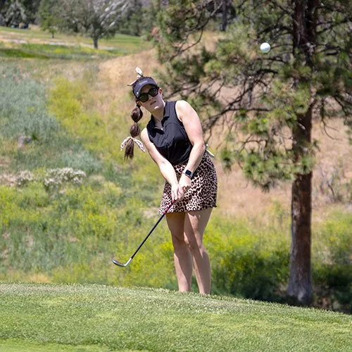 A woman playing golf at Fairview Mountain Golf Club with green grass and trees in the background, wearing sunglasses, a black sleeveless top, and a leopard-print skirt, swinging a golf club.