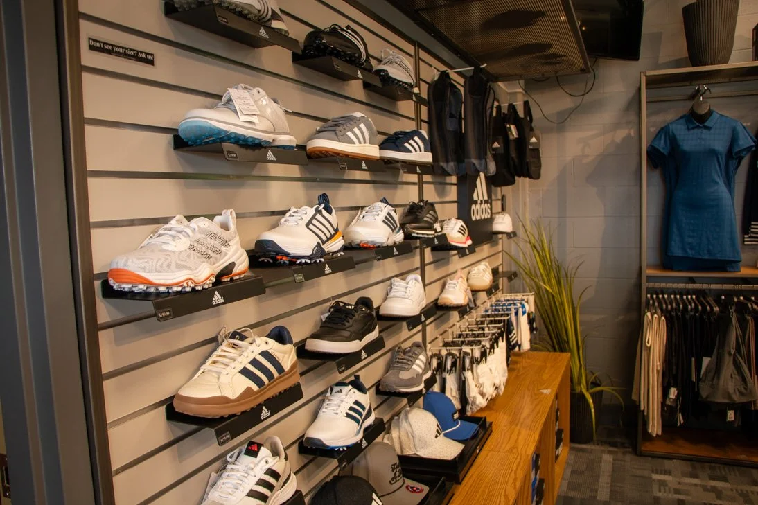 Display of various athletic shoes and hats on a wall-mounted rack inside a retail store, with clothing and accessories on a nearby shelf and a mannequin wearing a blue golf dress.