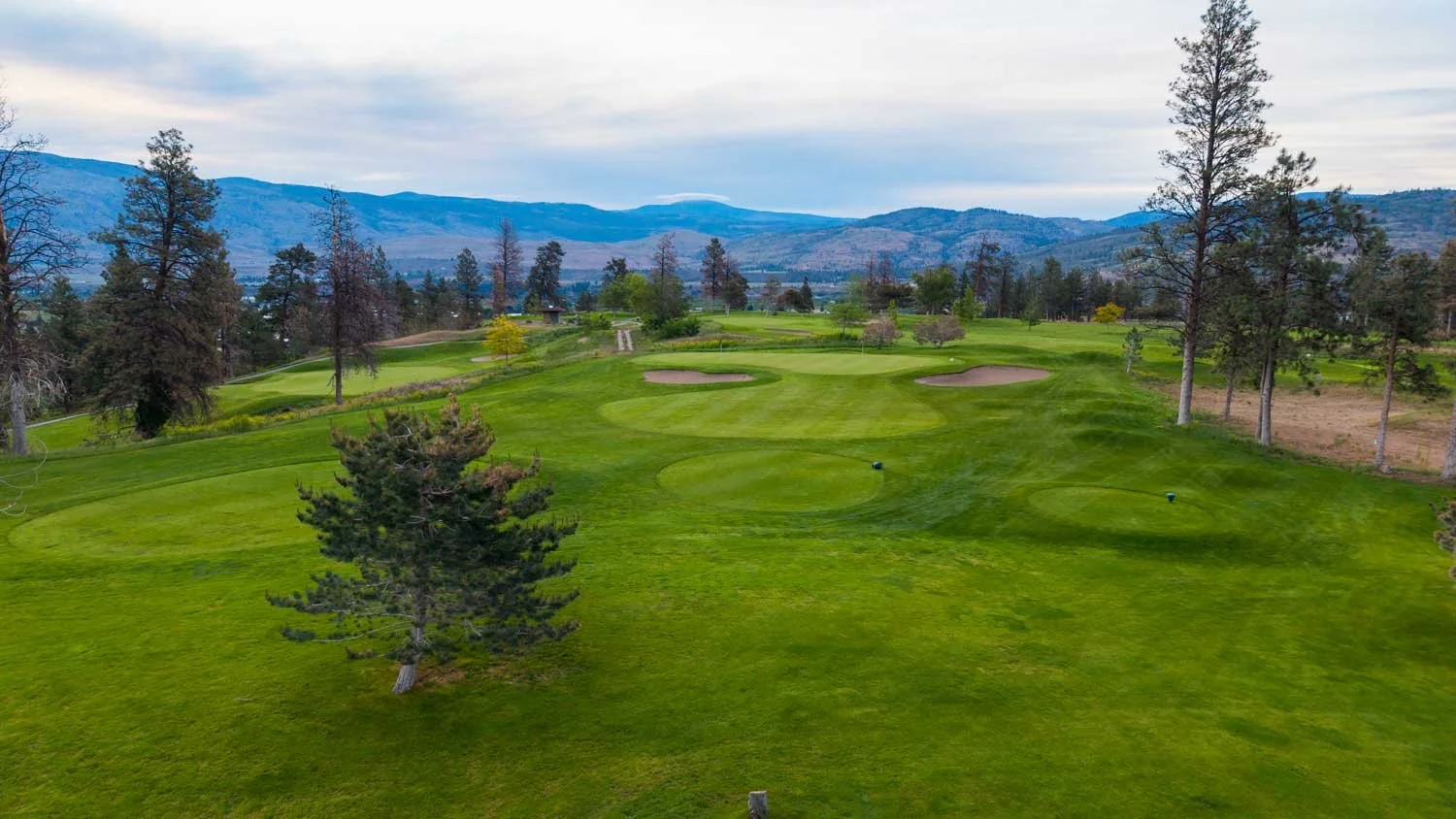 Scenic view of Fairview Mountain Golf Club golf course with green fairways, sand bunkers, and tall trees, set against a backdrop of mountains and a mostly cloudy sky.
