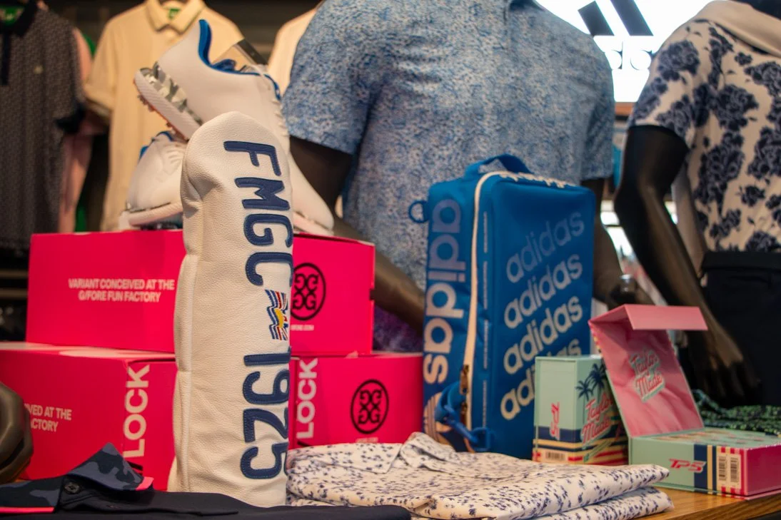 Display of golf accessories including golf club head covers, a pink box, a blue Adidas gym bag, and other golf-related items on the Fairview Mountain Golf Club Pro Shop store table.