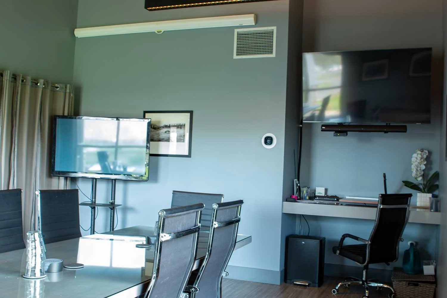 Fairview Mountain Golf Club meeting room with a glass-top table, black chairs, a wall-mounted TV, a smaller TV, a framed photograph, a white desk with office supplies, a black office chair, and decorative plants.