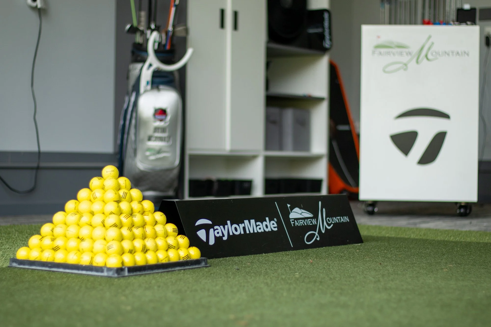 A display of yellow golf balls arranged on a black tray in front of a sign that reads 'TaylorMade' and 'Fairview Mountain.' In the background, there is a white storage unit with golf equipment and a white box with the Fairview Mountain logo.