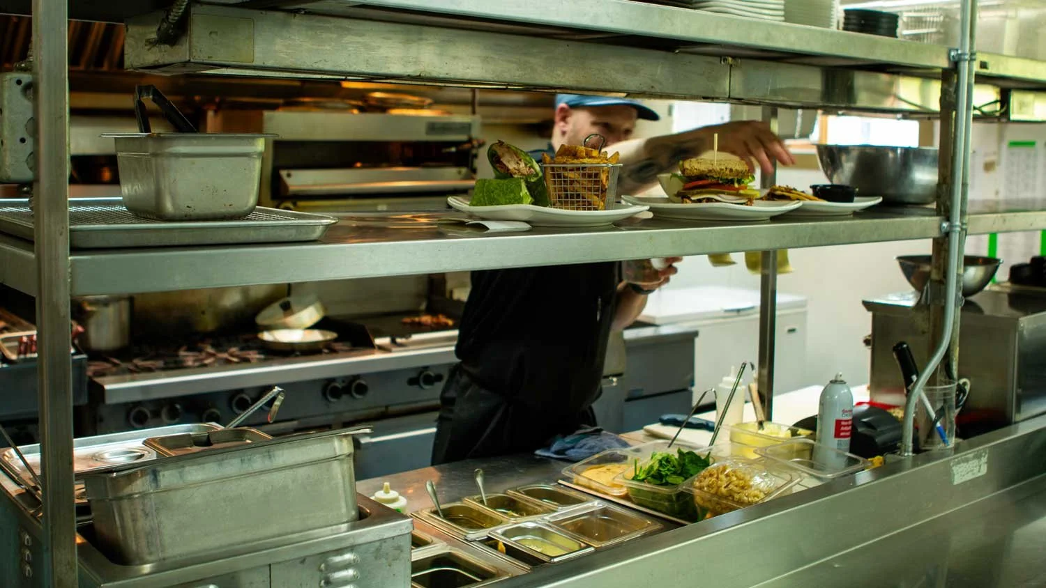 Behind-the-scenes view of the Fairview Mountain Golf Club restaurant kitchen with a chef placing plated dishes on a shelf, including hamburgers, fries, and greens, with various cooking utensils and ingredients on the counter.