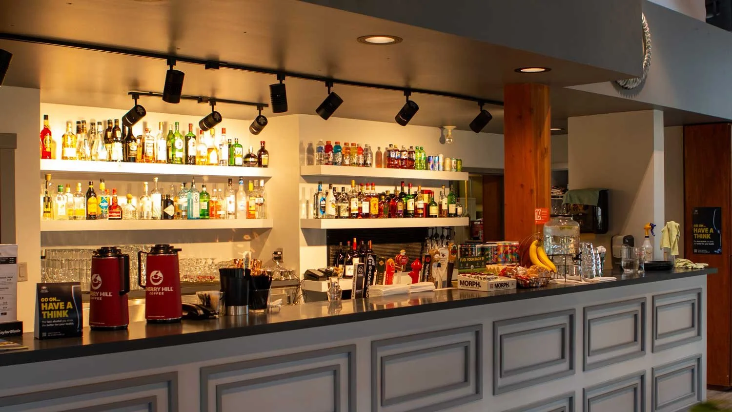 Fairview Mountain Golf Club Bar counter with bottles of alcohol on shelves, coffee dispensers, and snacks, in a modern lounge or bar area.