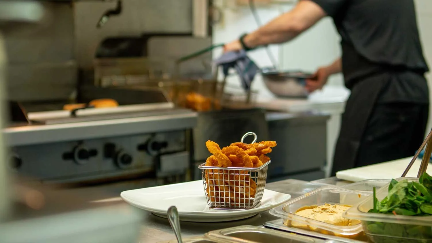 Basket of fried chicken tenders on a white plate in a restaurant kitchen with a chef preparing food in the background.