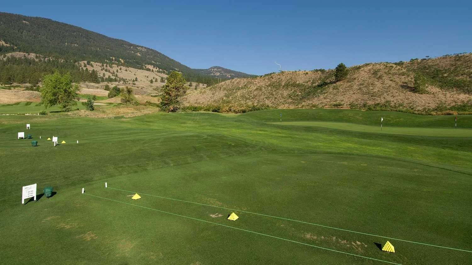 Fairview Mountain Golf Club driving range with markers and nets in a green landscape, mountains in the background, under a clear blue sky.