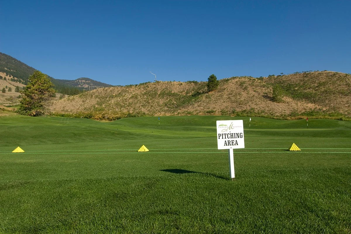 Fairview Mountain Golf Club practice area with a sign that reads "Pitching Area," on a grassy field under a clear blue sky, with mountains in the background.