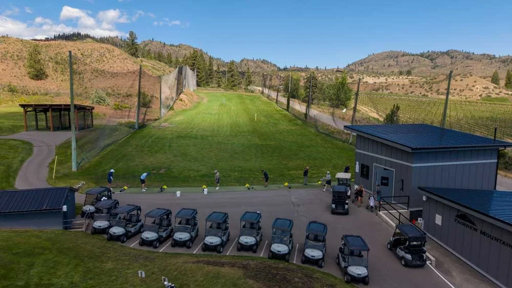 Fairview Mountain Golf Club driving range with golfers practicing, cars parked nearby, and hills in the background.