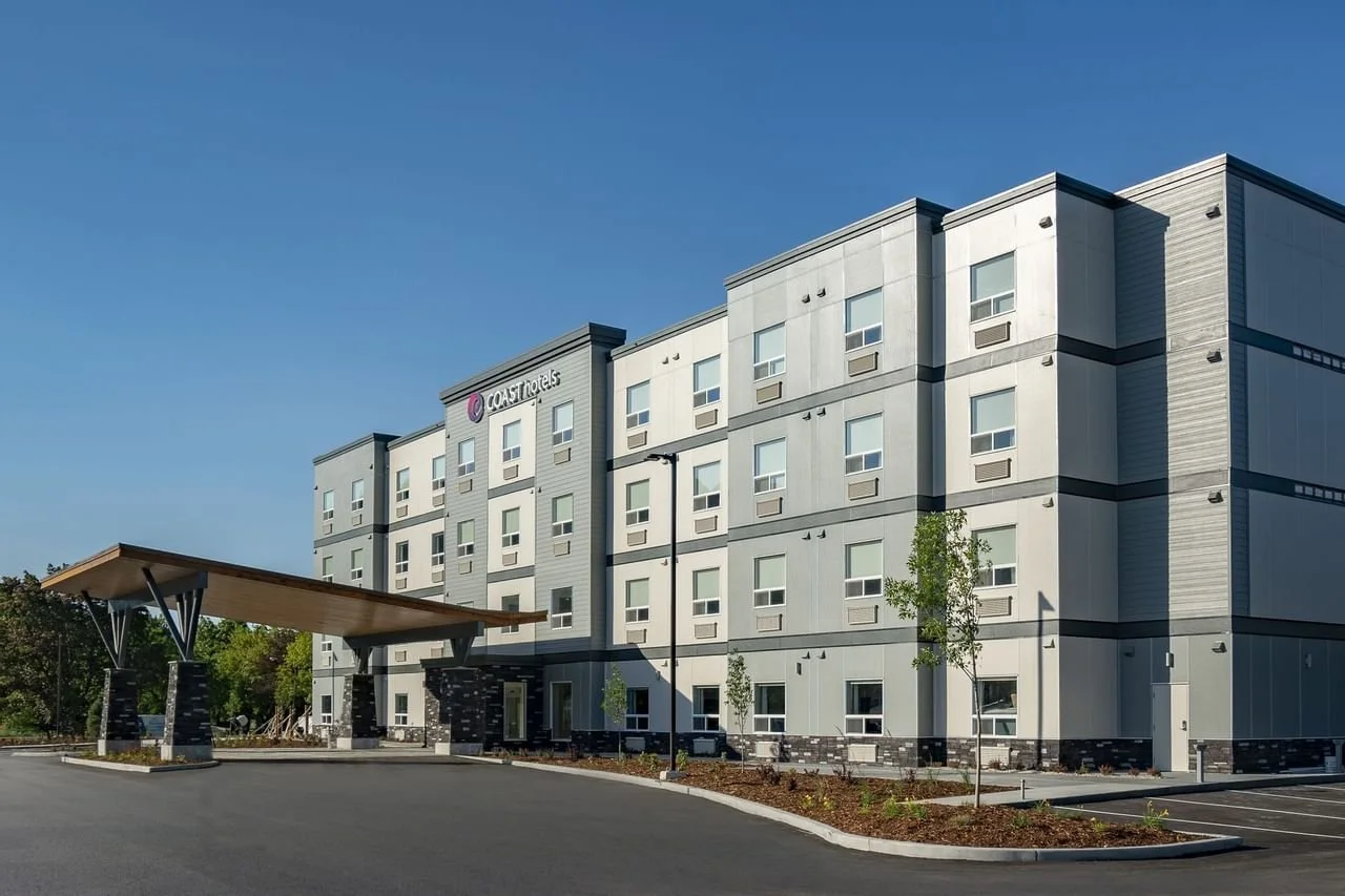 Exterior view of a modern, multi-story Coast Hotels building with a covered entrance and parking lot, surrounded by small trees and landscaping, under a clear blue sky.