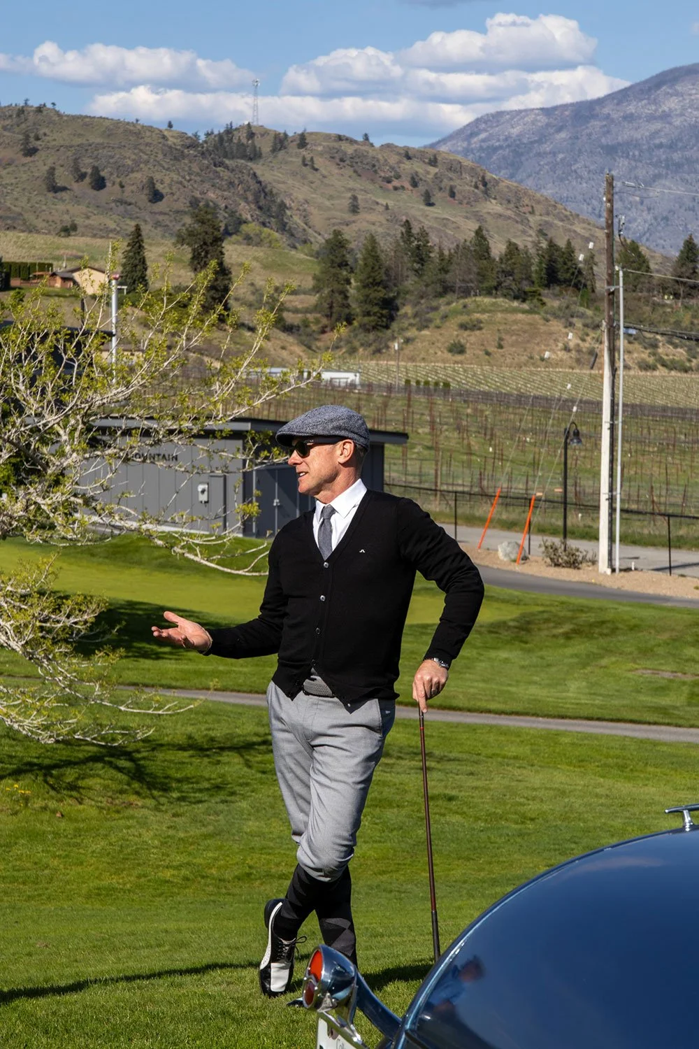 A man wearing a gray flat cap, sunglasses, a black sweater over a white shirt, gray pants, and golf shoes stands on a golf course, holding a golf club in his right hand and gesturing with his left hand while talking. Mountains and a blue sky with white clouds are visible in the background.
