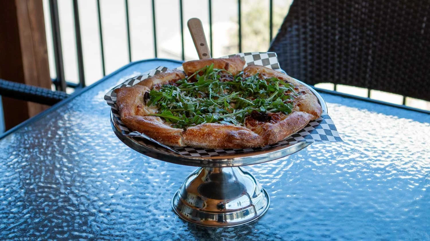 Pizza with arugula on a metal cake stand on a glass outdoor table.
