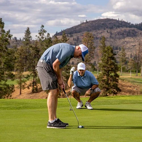 Two men playing Fairview Mountain Golf Club on a green with trees and mountains in the background, one preparing to putt and the other observing.