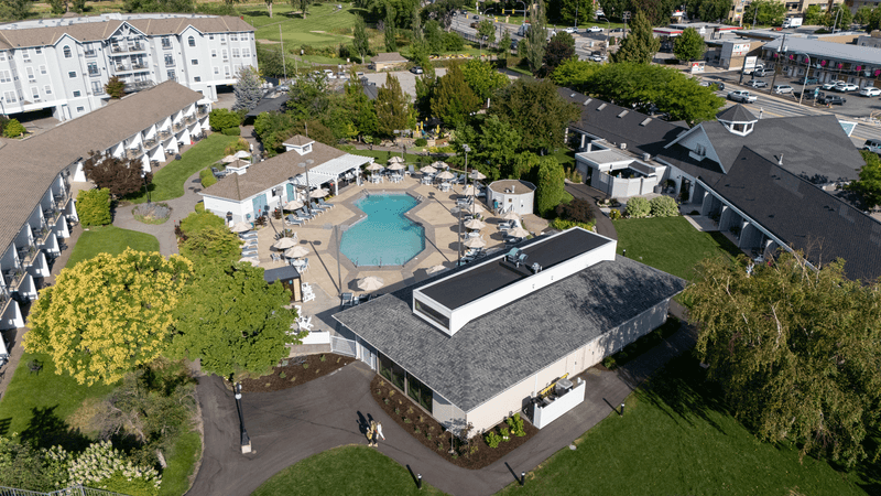 Aerial view of a residential apartment complex with a central swimming pool area, surrounded by lounge chairs and umbrellas, and adjacent building with a dark roof.