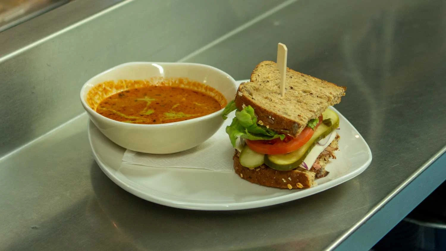 A bowl of tomato-based soup next to a veggie sandwich with cucumbers, tomato, lettuce, and mayonnaise on multigrain bread, served on a white plate on a metallic counter.