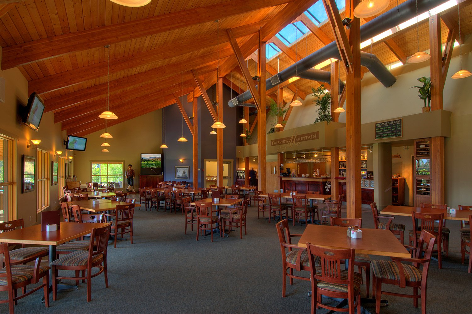 Interior of the Fairview Mountain Golf Club restaurant with wooden ceiling beams, multiple tables and chairs, large windows, and several TV screens on the walls.