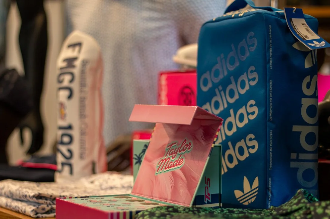 Close-up of an open pink TaylorMade box with golf tees, behind a blue Adidas backpack, on a table in Fairview Mountain Golf Club Pro shop.