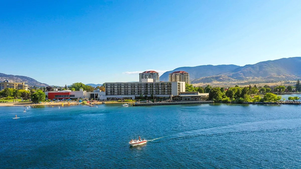 A scenic view of a large lake with boats, bordered by trees and modern buildings, with mountains in the background under a clear blue sky.