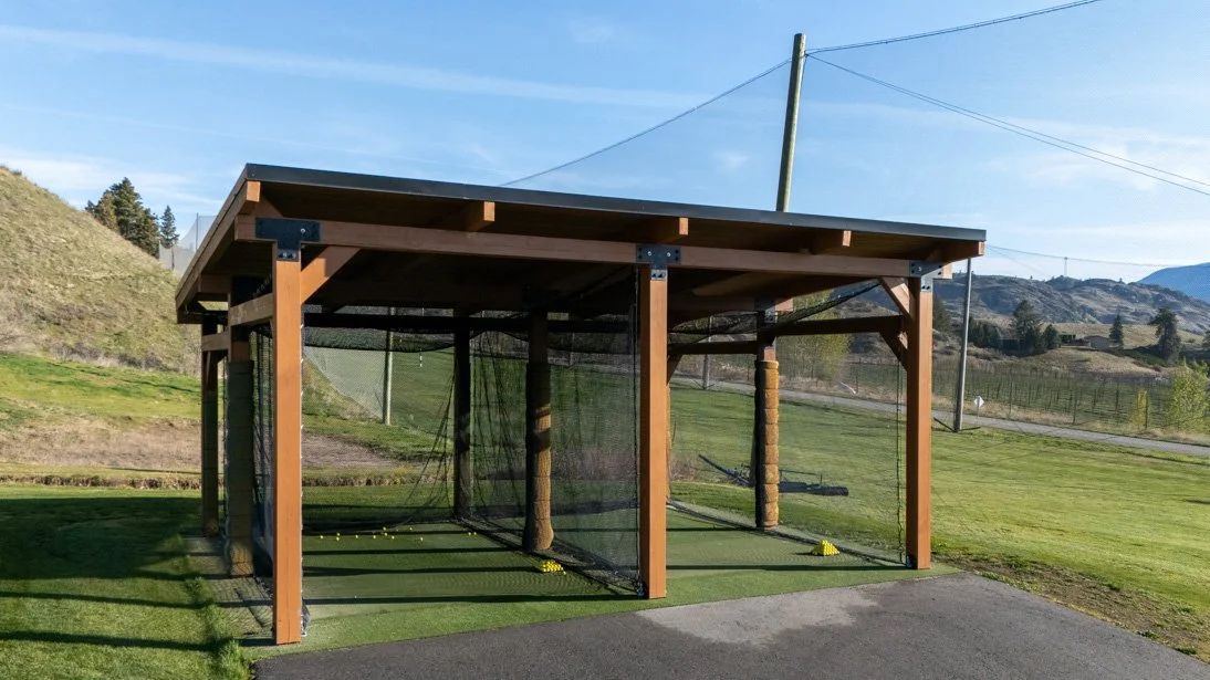 A wooden outdoor batting cage with netting, situated on a grassy field with rolling hills and mountains in the background.
