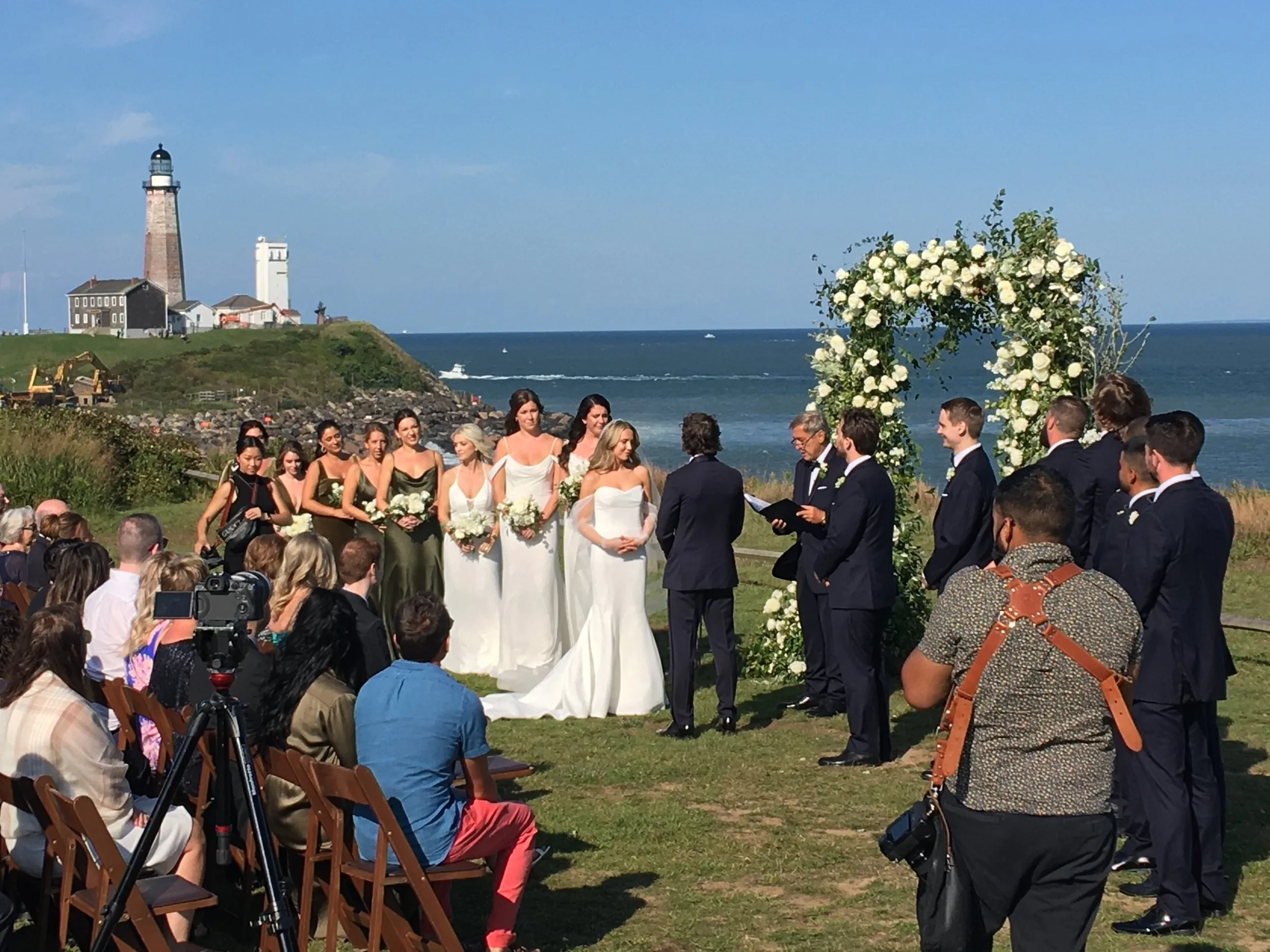 A wedding ceremony takes place outdoors near the coast, with a lighthouse and ocean in the background. The bride and groom stand under a floral arch, surrounded by bridesmaids and groomsmen, with seated guests watching.