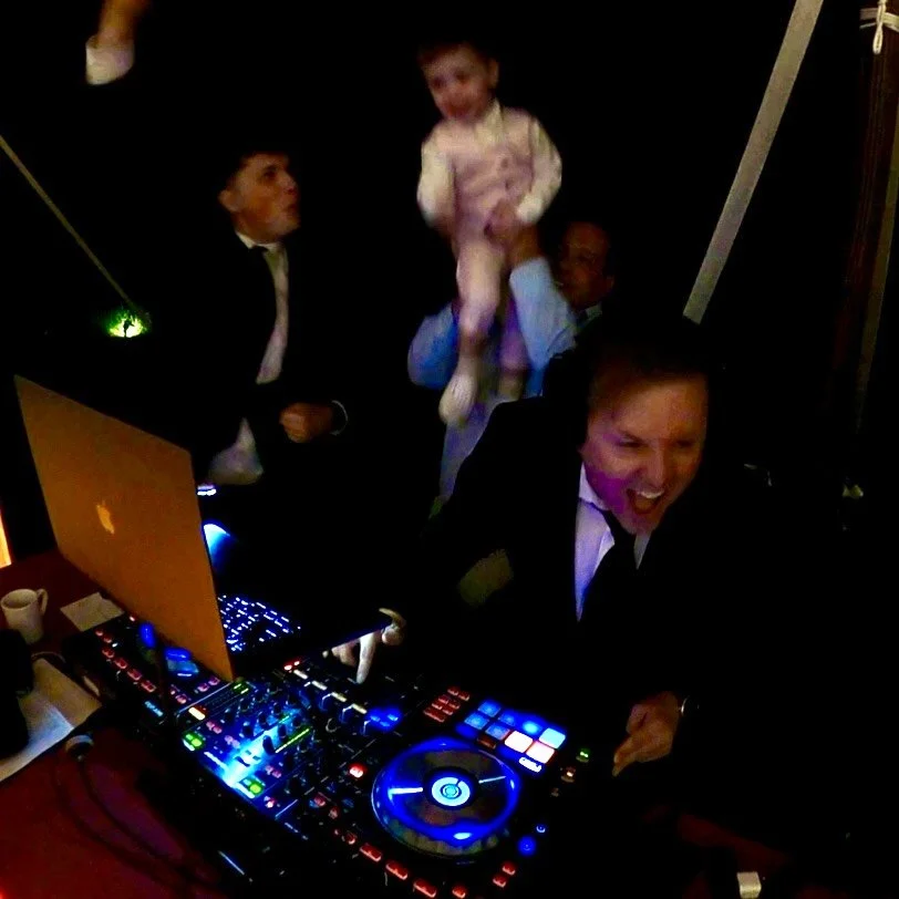 People dancing and celebrating at a party with DJ equipment, including a laptop and turntables.
