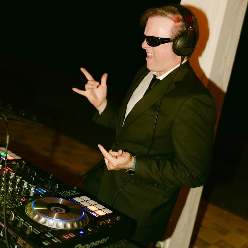 A man in a black suit and sunglasses wearing headphones, standing in front of DJ equipment, making rock gestures with his hands, at a party or concert.