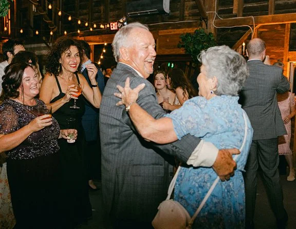 An elderly couple dancing together at a social gathering, with several women in the background holding drinks and smiling.