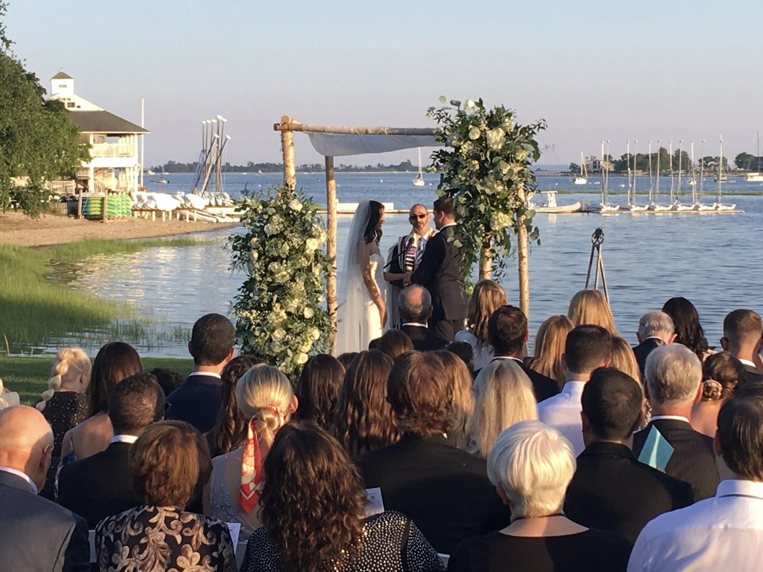 A wedding ceremony taking place by the water with a floral arch, a bride and groom, an officiant, and a seated audience, with sailboats on a lake in the background during sunset.
