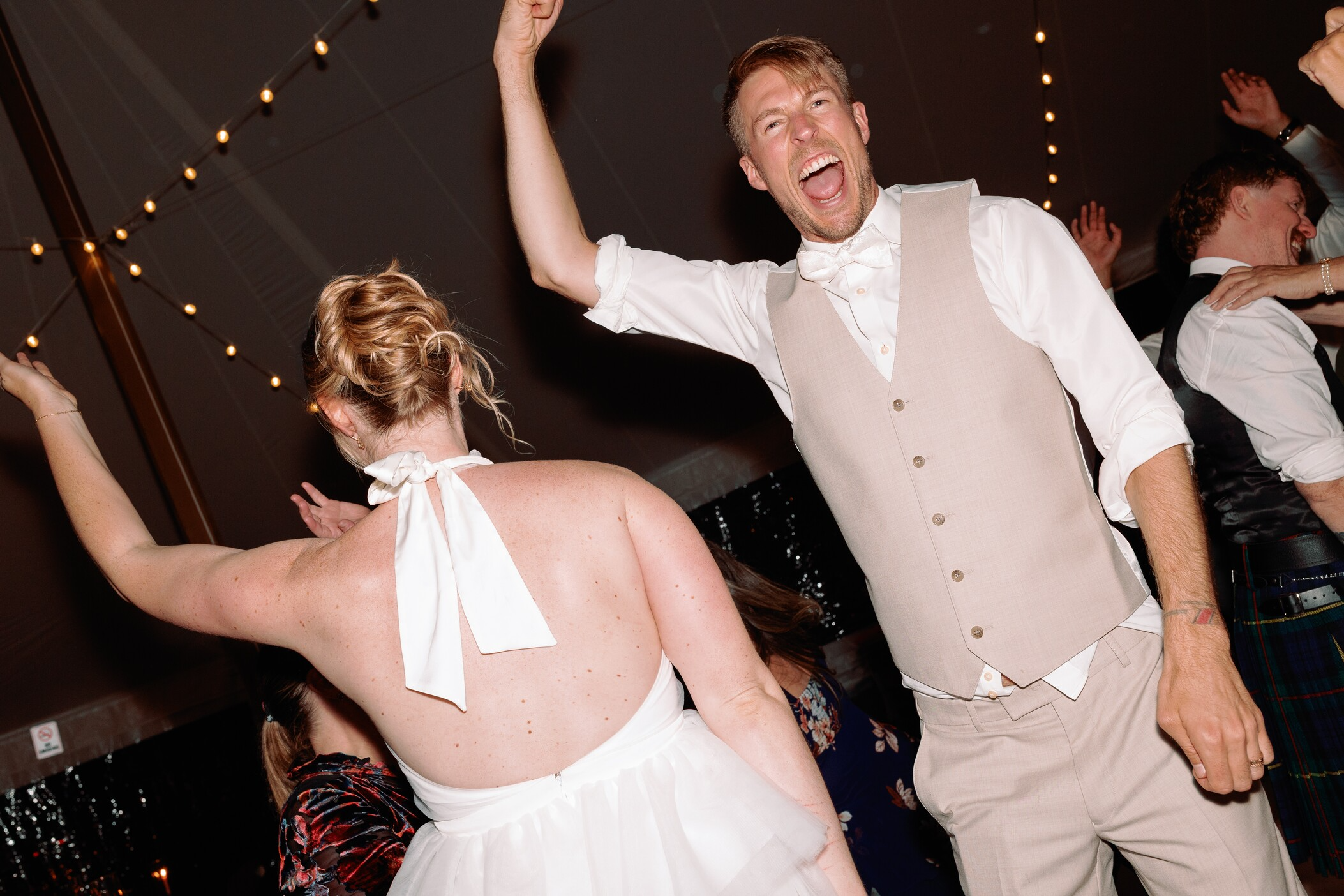 People dancing at a wedding reception, including a man in a beige vest and a woman in a white dress with a halter top, celebrating joyfully.
