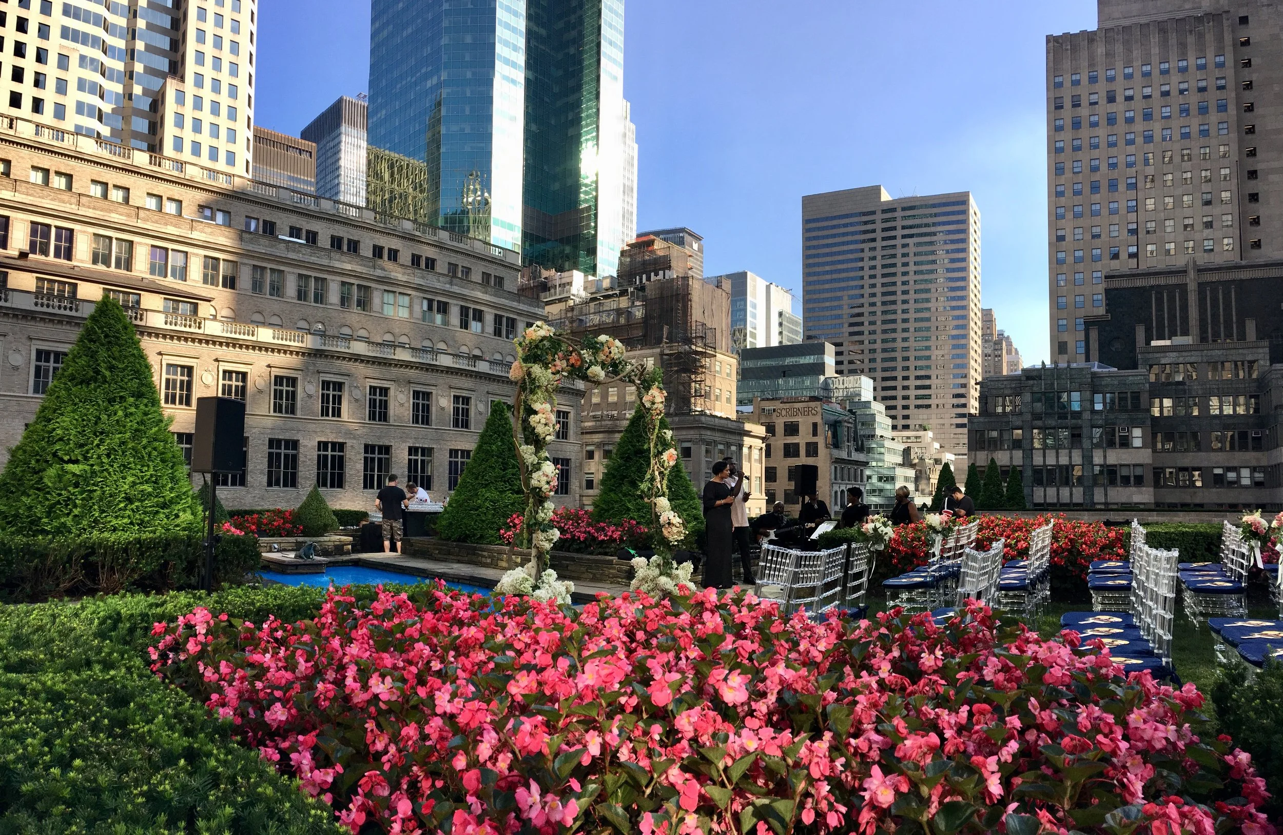 An outdoor wedding setup in a city rooftop garden with pink and white flowers, green bushes, chairs, and floral arch, surrounded by tall skyscrapers.